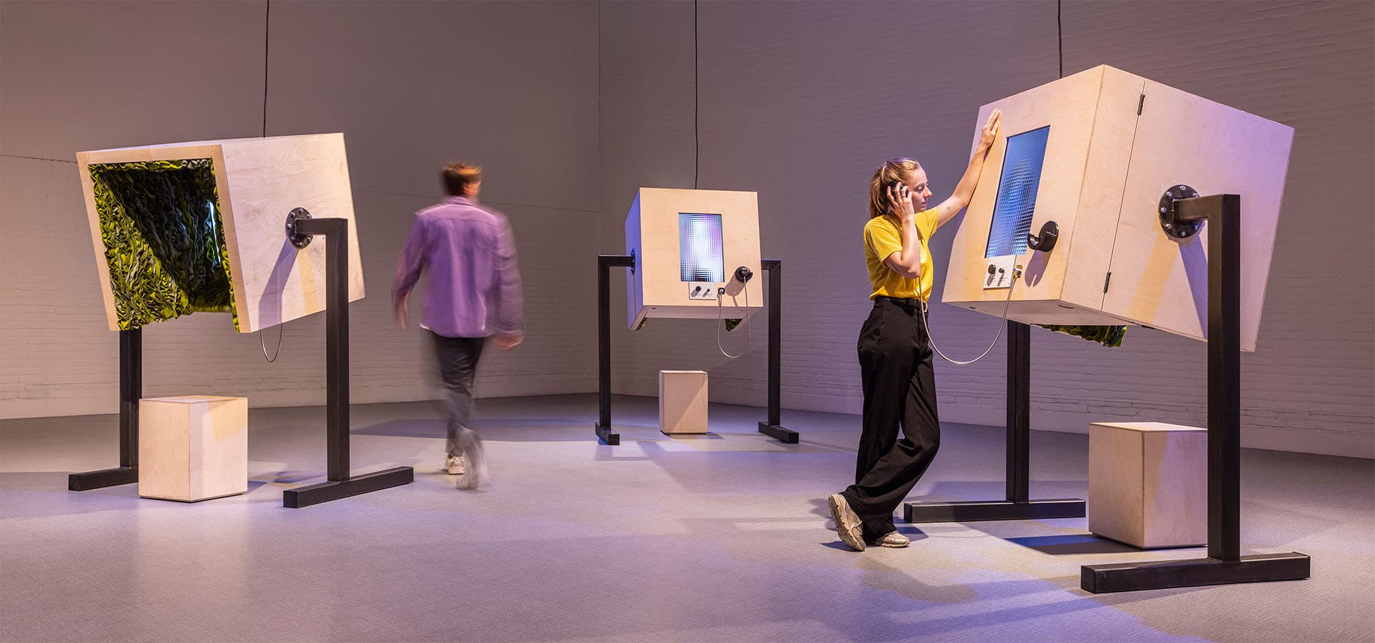 A visitor is attentively listening to past participants, while another is walking around the three objects that make up Channels.