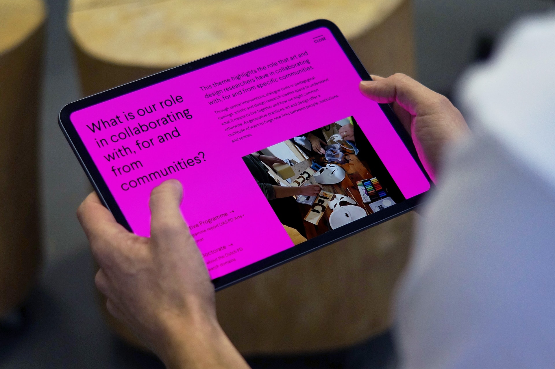 Two hands holding an iPad. The PD Arts+Creative website is displayed.